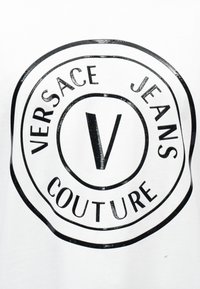 Μαύρο κυκλικό λογότυπο με το γράμμα "V" στο κέντρο και τις λέξεις "Versace Jeans Couture" γύρω του σε λευκό φόντο.