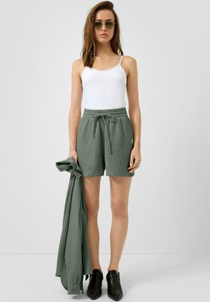 Kobieta ubrana w biały top na ramiączkach, zielone krótkie spodenki ze sznurkiem, ciemne botki za kostkę, okulary przeciwsłoneczne, trzymająca dopasowaną zieloną kurtkę w prawej ręce.