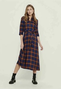 Vestido de camisa a cuadros en marrón y azul, con botones en la parte delantera, cintura ceñida con cinturón y mangas de tres cuartos, combinado con botas negras hasta el tobillo.
