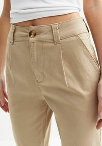 Beige Hose mit glatter Textur, ausgestattet mit einem Knopfverschluss, seitlichen Taschen und Gürtelschlaufen. Das Design umfasst eine lockere Passform.