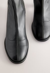 Sorte læderankelstøvler med en glat finish, der har en sidesyning og kontrasterende syninger langs sømmene. Runde tæer og flade såler.