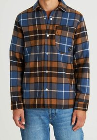 Koszula z flaneli w niebiesko-brązowo-czarną kratę z białymi liniami, z pojedynczą kieszenią na piersi i zatrzaskami. Noszona z jasnoniebieskimi dżinsami.