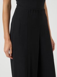 Κοντινό πλάνο μαύρων ψηλόμεσων παντελονιών με πλαϊνές τσέπες, φορεμένα με μια τυλιγμένη μαύρη μπλούζα, εμφανίζοντας τα χέρια του μοντέλου από τις πλευρές.