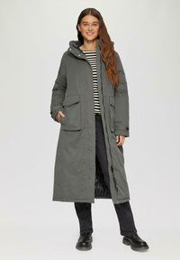 Manteau long grey matelassé avec capuche, deux poches avant, fermeture éclair et doublure chaude. Porté par-dessus une chemise à rayures noir et blanc.