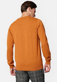 Pomarańczowy sweter wykonany na drutach z długimi rękawami, ściągaczami przy nadgarstkach i u dołu, z okrągłym dekoltem. Nosi się go na szare w kratę spodnie.