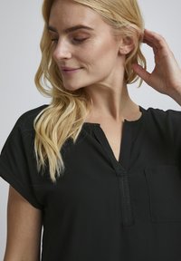 Schwarze Bluse mit rundem Ausschnitt, kurzen Ärmeln und einer Tasche auf der linken Seite. Der Stoff wirkt glatt mit einem subtilen Glanz.
