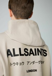 Sudadera con capucha beige con el texto impreso en negro "ALLSAINTS" y otros caracteres en la parte posterior. Tela suave, diseño casual, detalles de costura visibles.