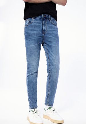 Personne portant un jean slim bleu, une chemise noire et des baskets blanches avec des touches vertes, debout les bras croisés devant un fond blanc.