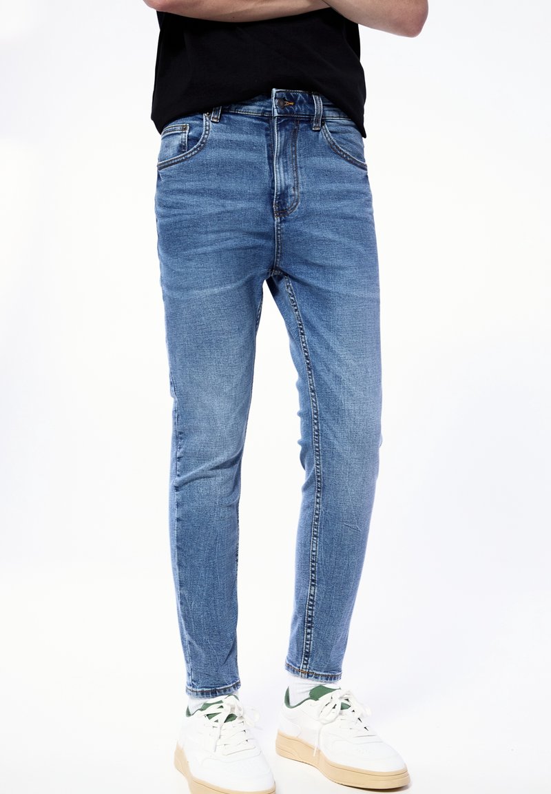 Personne portant un jean slim bleu, une chemise noire et des baskets blanches avec des touches vertes, debout les bras croisés devant un fond blanc.