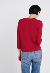 Czerwony prążkowany sweter z dzianiny z rękawami do łokcia, z wiązaniami po bokach. Noszony z jasnoniebieskimi jeansami z wysokim stanem. Widok od tyłu.