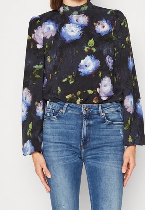 Femme portant un chemisier noir à manches longues avec un imprimé floral bleu et violet, rentré dans un jean bleu délavé.