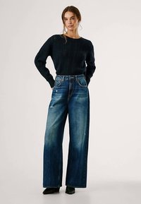 Dark blue knitted sweater with textured pattern, paired with high-waisted light blue distressed wide-leg jeans. Model stands in neutral background.