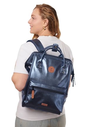 Junge Frau trägt weißes Hemd und Jeans und hat einen glänzenden dunkelblauen Rucksack mit vorderer Reißverschlusstasche und Leder-Logopatch.