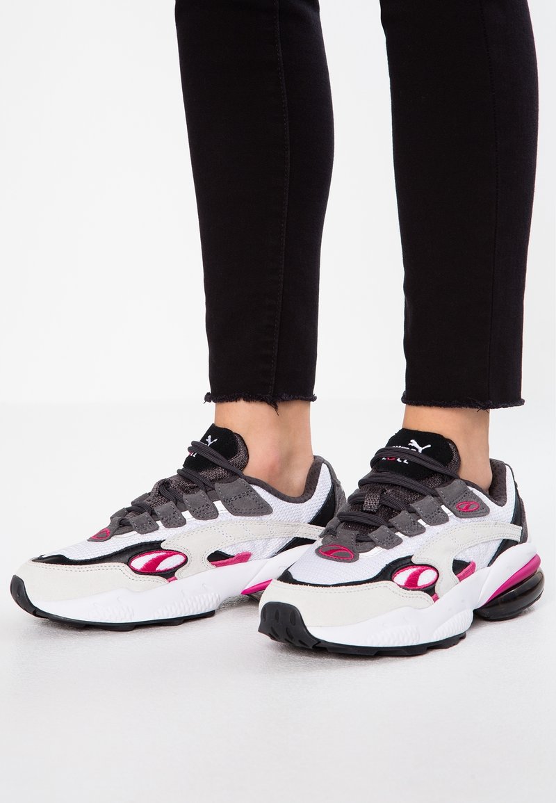 Les baskets présentent un mélange de matériaux blancs, gris et noirs avec des accents roses. Les textures en maille et en daim rehaussent le design. Semelle épaisse.