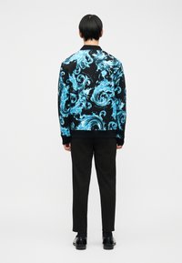 Άτομο που φοράει μαύρο παντελόνι και μαύρα παπούτσια, με μαύρο και μπλε πουκαμίσα με μακριά μανίκια με σχέδιο, στέκεται με την πλάτη προς τα εμπρός σε λευκό φόντο.