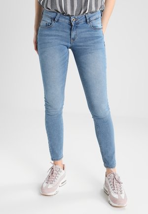 Kobieta w jasnoniebieskich obcisłych dżinsach i białoróżowych sneakersach, stojąca z rękami w kieszeniach na jednolitym tle.