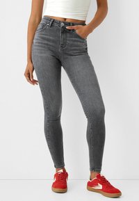 Szürke, magas derekú farmernadrág, testhezálló kialakítással, öt zsebbel és sima, enyhén texturált denim anyagból, piros sportcipővel párosítva.