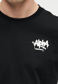 Άνδρας που φοράει μαύρη μπλούζα με μικρό λευκό λογότυπο "Alpha Industries" στο αριστερό στήθος.