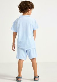 Lysblå bomulds polo shirt med korte ærmer og shorts sæt, blød tekstur, med klassisk krave og afslappet pasform, parret med mørke sandaler.