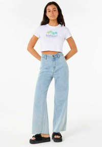 Camiseta blanca corta con gráfico de palmera y jeans de pierna ancha de talle alto en azul. La modelo lleva sandalias de plataforma negras. Fondo liso.