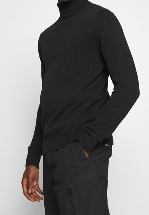 Homme portant un pull à col roulé noir à manches longues et un pantalon cargo noir, une main dans la poche, sur fond uni.