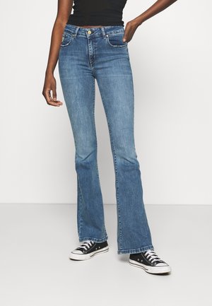 Kvinde iført blå bootcut jeans, sort tanktop og sorte lave sneakers står med den ene hånd i lommen.