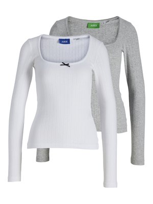 Femme debout, portant un haut à manches longues gris clair, un pantalon cargo ample vert olive et des bottes noires mi-mollet sur fond blanc.