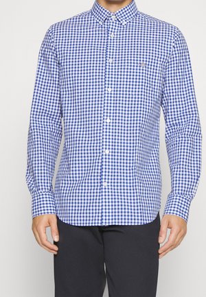 Uomo che indossa una camicia a quadri blu e bianca a maniche lunghe con pantaloni neri su uno sfondo semplice.