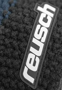 Black textured fabric featuring a prominent white "reusch" logo on a black leather patch, with a soft, fuzzy surface detail.