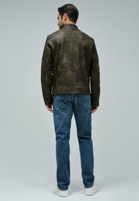 Giacca in pelle marrone con finitura testurizzata, colletto rialzato e polsini con zip, abbinata a jeans blu e sneakers bianche.