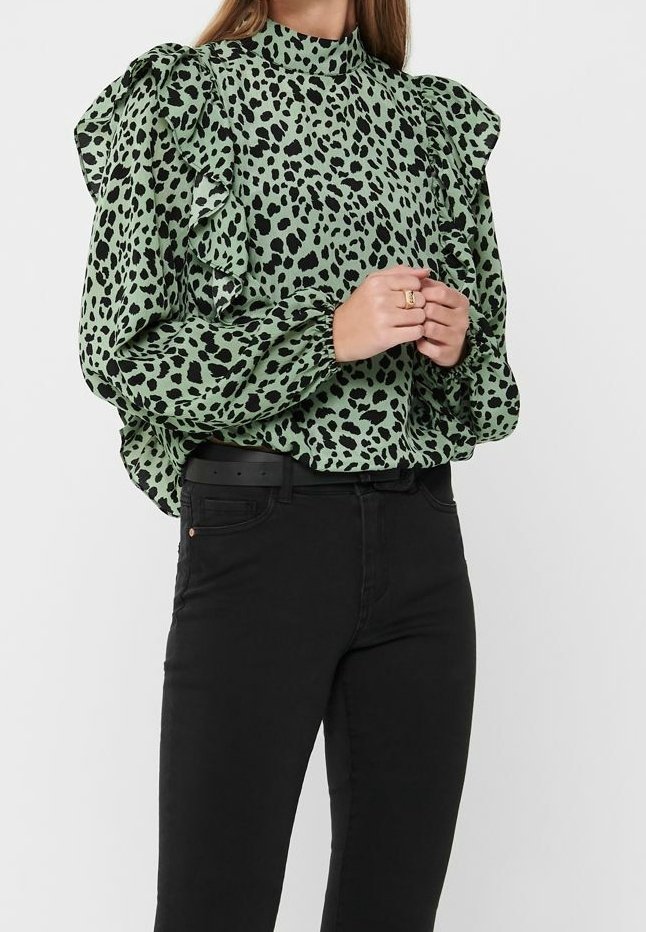 Femme portant un chemisier vert à manches longues avec des taches léopard noires et des volants, associé à un pantalon noir taille haute et une ceinture noire.