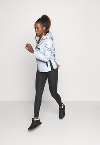 Hellgraue, kapuzenjacke mit einem Frontreißverschluss und schwarzem Logo; kombiniert mit schwarzen, strukturierten Leggings und schwarzen Turnschuhen. Glatte Stoffoberfläche.