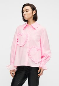 Γυναίκα που φοράει ανοιχτό ροζ πουκάμισο με κουμπιά, με βολάν σε σχήμα καρδιάς και ρεβέρ με βολάν, συνδυασμένο με μαύρο δερμάτινο παντελόνι.