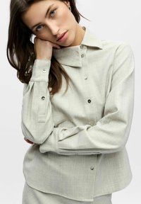 Jeune femme aux yeux bleus reposant sa tête sur sa main, portant une chemise gris clair boutonnée avec des boutons en métal, sur un fond blanc.