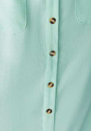 Vaaleanvihreä nappikauluspaita, jossa on sileä rakenne, keskellä ruskeat napit ja kaksi rintataskua.