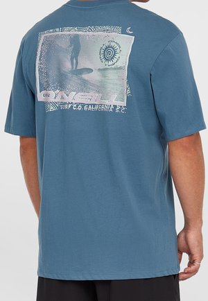 Homme portant un t-shirt bleu avec un motif de surf délavé et le texte "O'Neill Surf Co. California S.C." au dos.