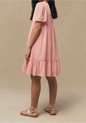 Fille portant une robe rose texturée à manches bouffantes, longueur genou, et des sandales argentées, debout de profil sur un fond neutre.