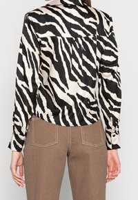 Blusa di satin con un motivo a strisce zebra in bianco e nero, dotata di colletto, polsini bottonati e orlo arrotondato. Il retro presenta un dettaglio plissettato.