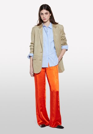 Blazer beige in camoscio, camicia a righe blu chiaro e pantaloni larghi arancione lucido. Le scarpe basse completano l'outfit, con texture morbide e colori vivaci.