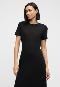 Μαύρο βαμβακερό φόρεμα t-shirt με κοντά μανίκια, στρογγυλή λαιμόκοψη και εφαρμοστή σιλουέτα. Η ραφή στο μπροστινό μέρος προσθέτει ορισμό.