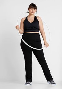 Čierny športový crop top s prekríženým dizajnom, v kombinácii s čiernymi legínami. Modelka drží biely hula hoop a má na nohách sivé tenisky.