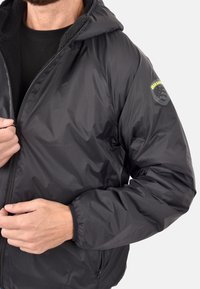 Giacca nera isolata con cappuccio, presenta una superficie esterna liscia, un interno foderato in pile, polsini elasticizzati e un logo sulla manica.