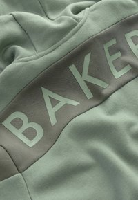 Mintová mikina s hladkou texturou, která má tmavě šedou část a vyvýšené písmo se slovem "BAKER" v světle zelené barvě na přední straně.