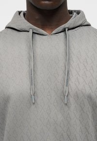 Κοντινό πλάνο γκρι φούτερ με διακριτικό γεωμετρικό μοτίβο και κορδόνια, φορεμένο από άτομο με σκούρο δέρμα.