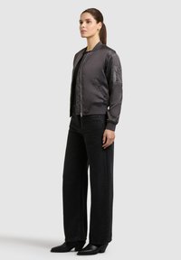 Femme debout portant une veste bomber gris foncé, un pantalon large noir, et des bottes à talons noires, sur un fond uni gris clair.