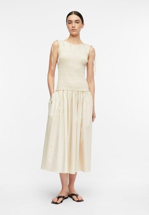 Frau, die ein ärmelloses, cremefarbenes Midikleid mit geraffter Taille und schwarzen flachen Sandalen vor einem schlichten weißen Hintergrund trägt.
