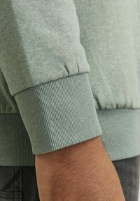 Sudadera verde claro con puños acanalados y una textura suave, mostrando un primer plano del área del brazo y la muñeca.