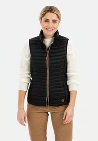Zwarte puffervest met een rits aan de voorkant, met een opstaande kraag, gestikte design en een leren rits met bruine kleur. Gedragen over een crèmekleurige trui.