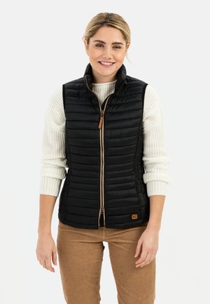 Zwarte puffervest met een rits aan de voorkant, met een opstaande kraag, gestikte design en een leren rits met bruine kleur. Gedragen over een crèmekleurige trui.