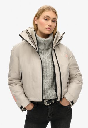 Beżowa ocieplana kurtka z wysokim kołnierzem, czarnym zamkiem błyskawicznym i detalem z logo. Noszona na szarym swetrze dzianinowym i czarnych dżinsach, z paskiem.